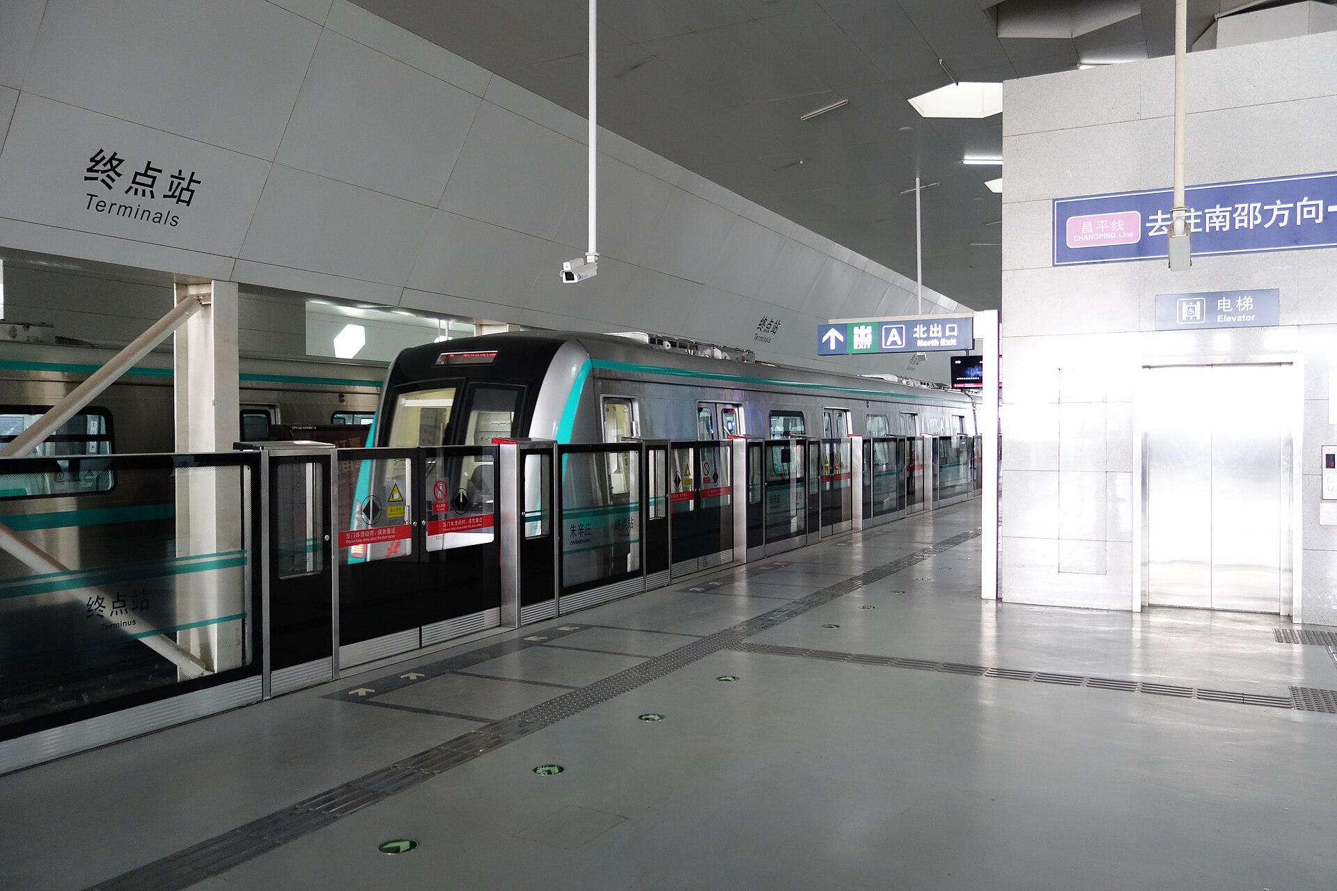 A turquoise-striped Beijing Subway Line 8 train stands at Zhuxinzhuang, beside platform barriers and bilingual exit signs.