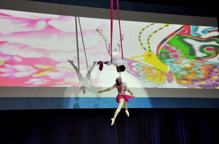 Two aerial acrobats perform above a Beijing stage, with one inverted in silks and the other suspended below.