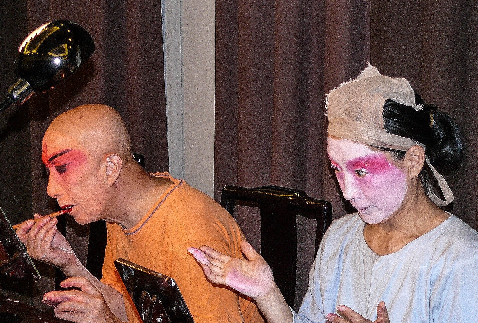 Two Peking Opera performers in Beijing apply bright face makeup at a dressing table using small handheld mirrors.