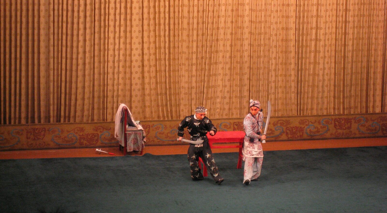 Two Peking Opera performers in Beijing hold stage weapons before a gold curtain, with audience silhouettes along the foreground.