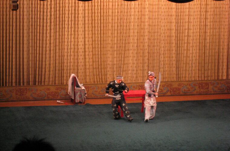 Two Peking Opera performers in Beijing hold stage weapons before a gold curtain, with audience silhouettes along the foreground.