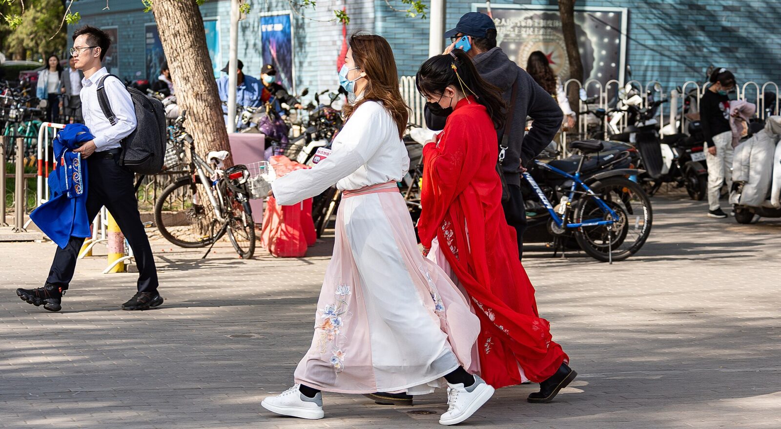 Two visitors wearing white and red hanfu walk past bicycles near the east gate of Beijing Yuyuantan Park.