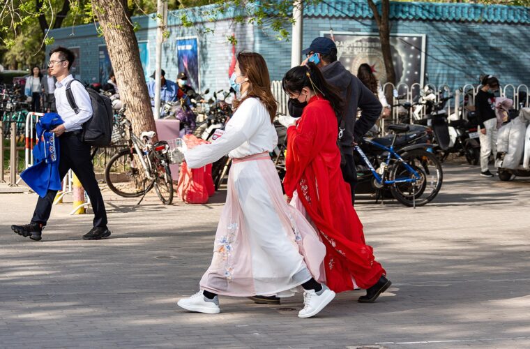 Two visitors wearing white and red hanfu walk past bicycles near the east gate of Beijing Yuyuantan Park.