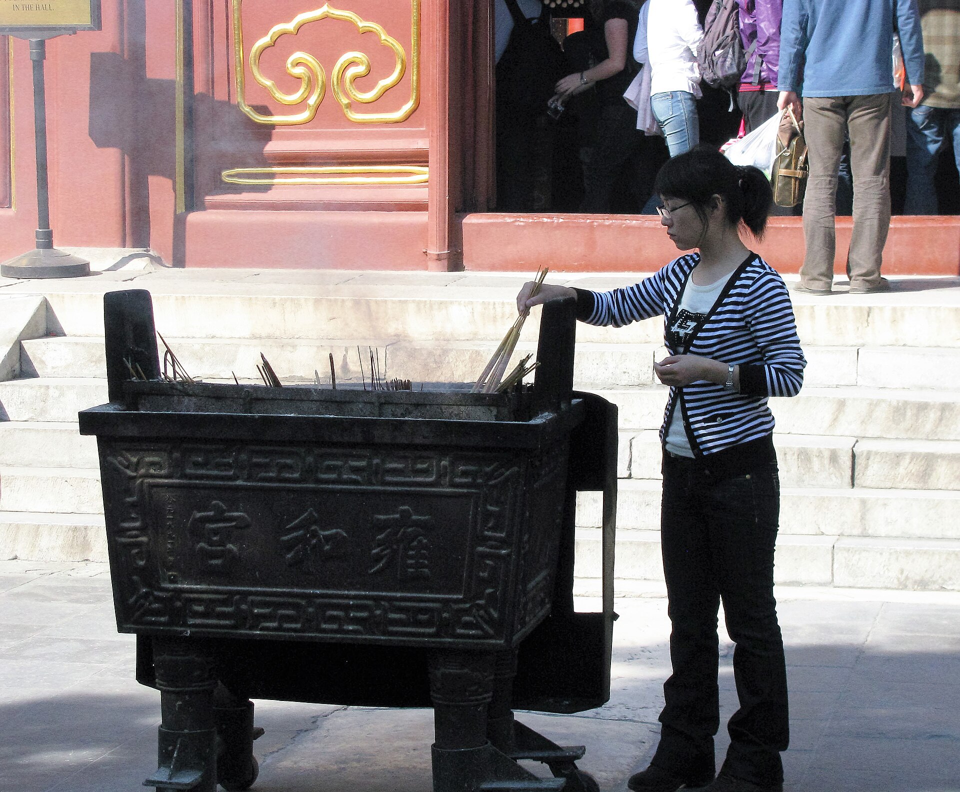 A visitor places incense sticks into a large metal burner beside red doors and stone steps at Yonghe Temple in Beijing.