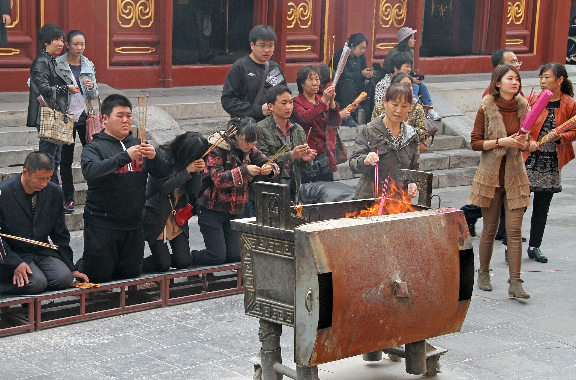Visitors at Beijing Yonghe Temple hold incense sticks and bow beside a smoking burner in the paved courtyard.
