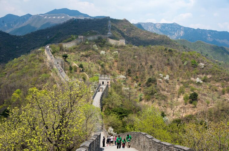Visitors climb the Great Wall at Mutianyu as watchtowers trace wooded ridges beneath distant rocky mountains.