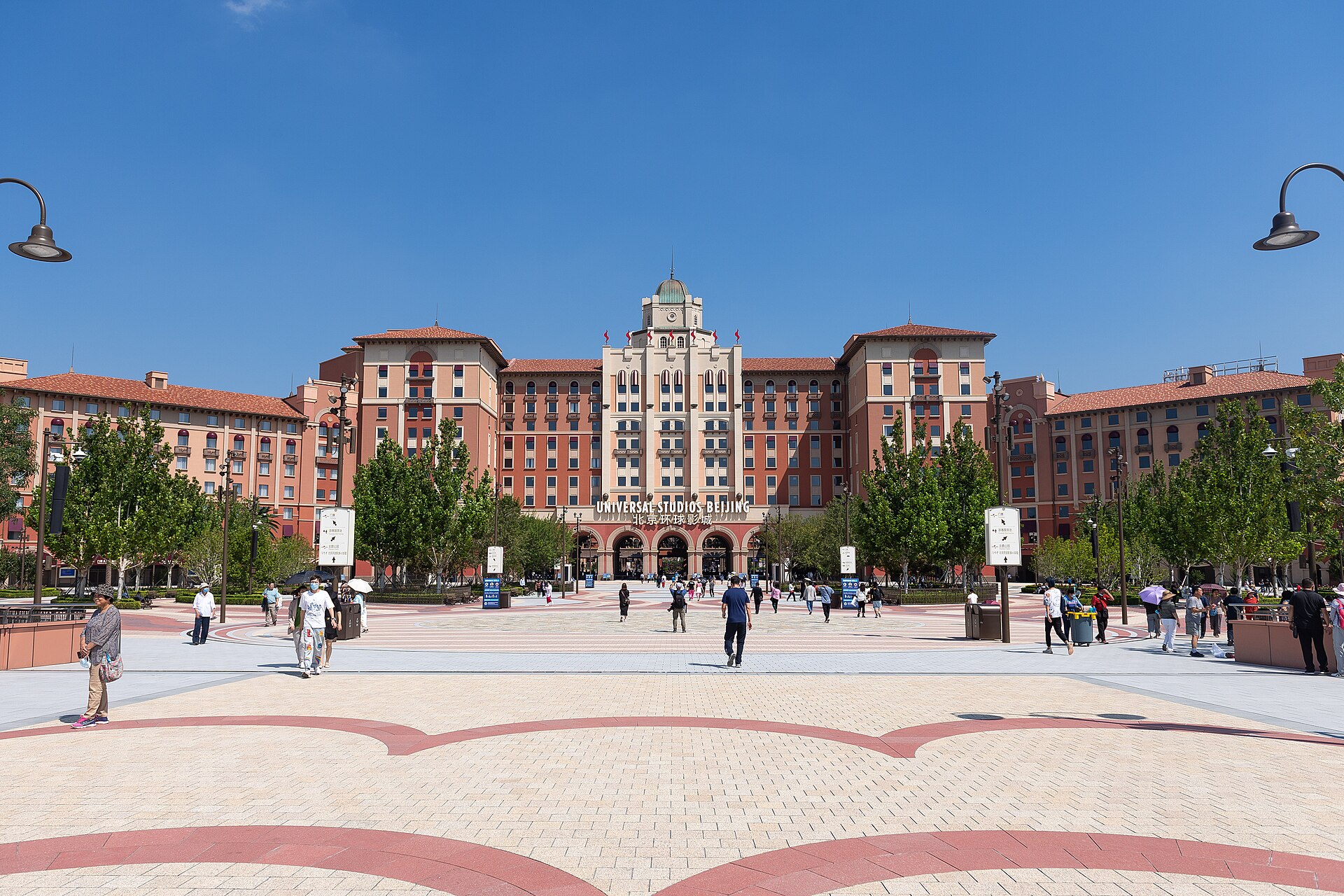 Visitors cross the plaza toward Universal Studios Beijing's entrance, set beneath the central arches of a large symmetrical hotel building.