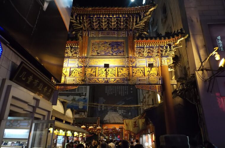 Visitors crowd beneath an illuminated gateway and red lanterns at Wangfujing Snack Street in Beijing after dark.