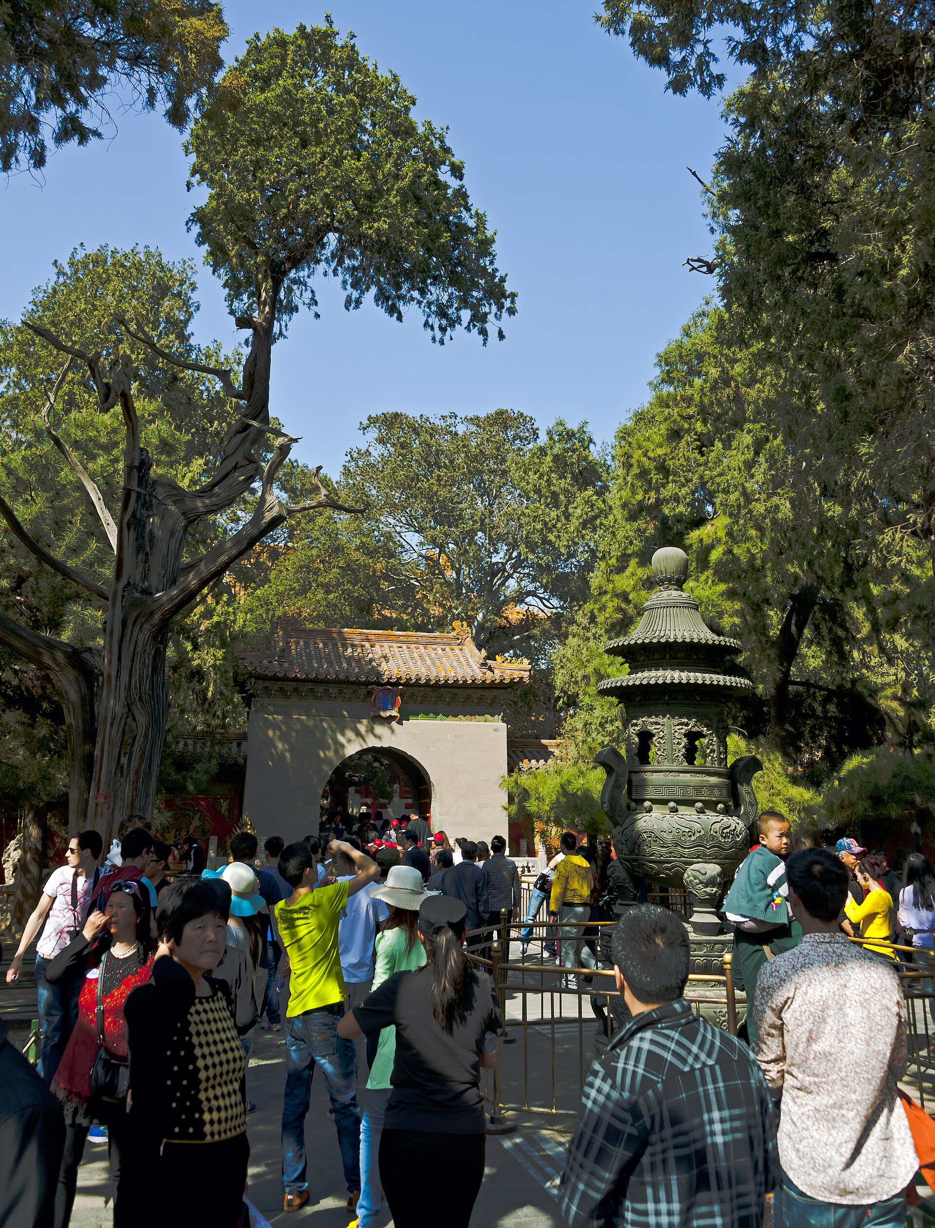 Visitors crowd the Forbidden City's Imperial Garden beside a bronze incense burner, with a child riding on an adult's shoulders.