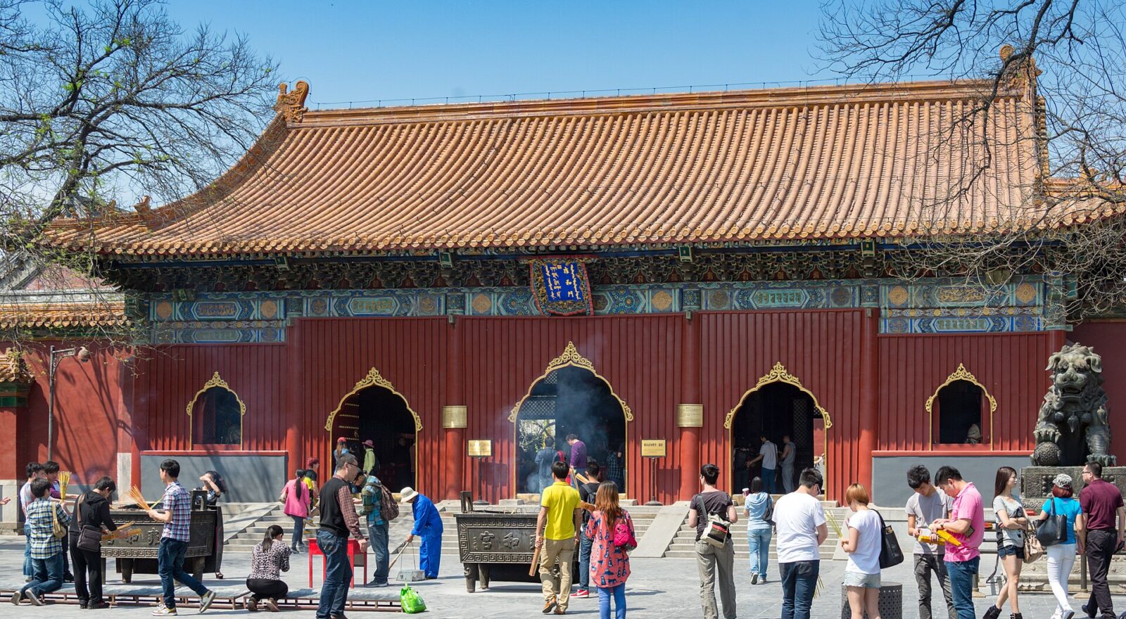 Visitors gather around incense burners before a red, yellow-roofed hall at Beijing's Lama Temple under a clear blue sky.