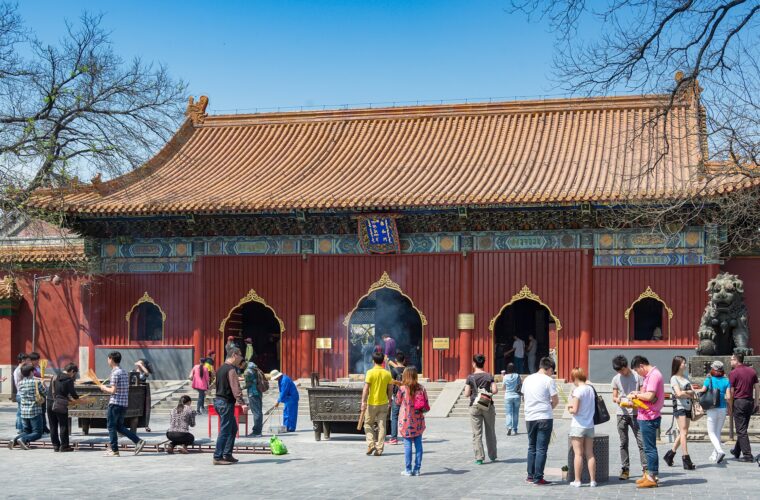 Visitors gather around incense burners before a red, yellow-roofed hall at Beijing's Lama Temple under a clear blue sky.