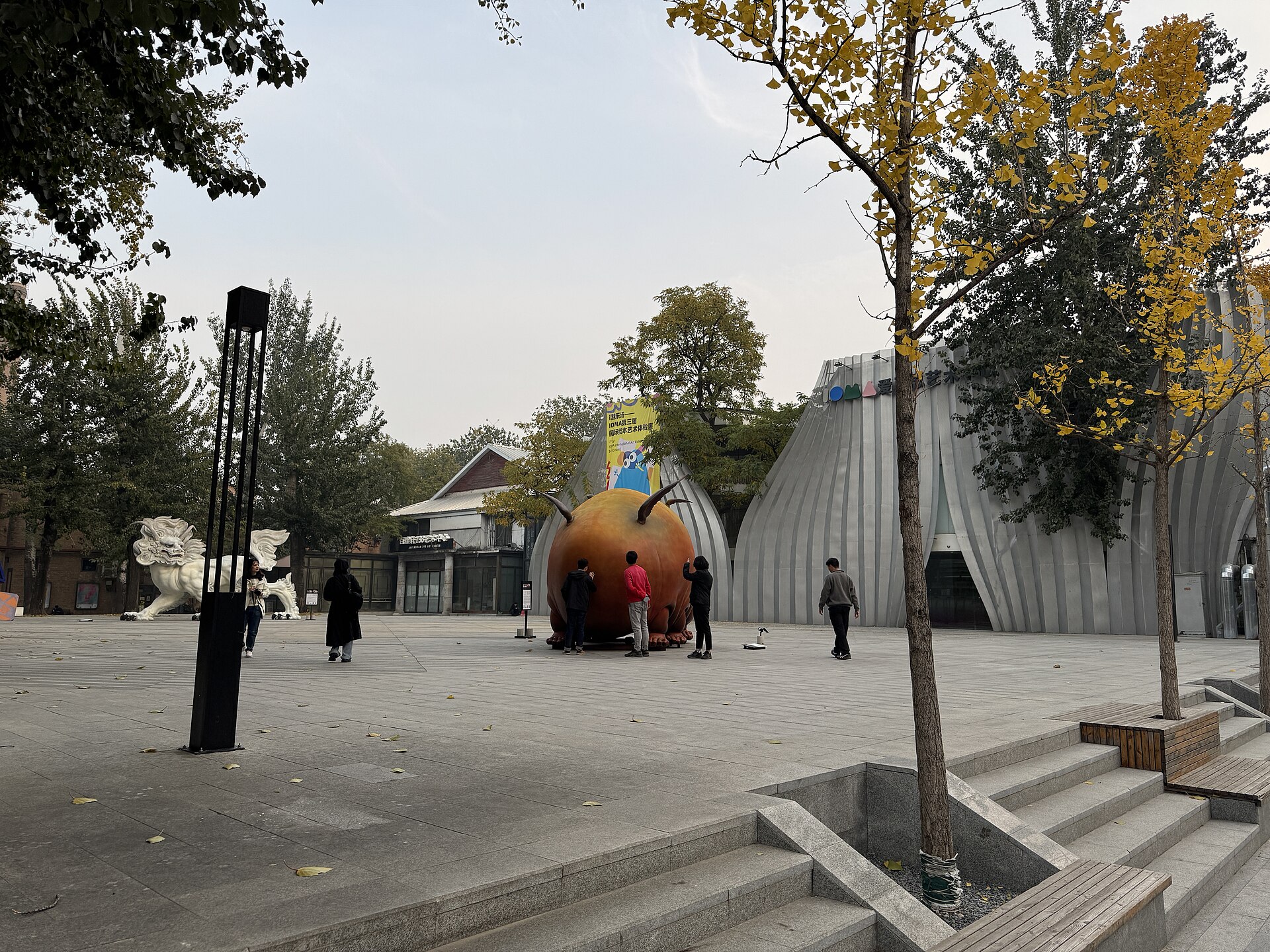 Visitors gather around a large orange sculpture beside a silver gallery building and autumn trees in Beijing's 798 Art District.