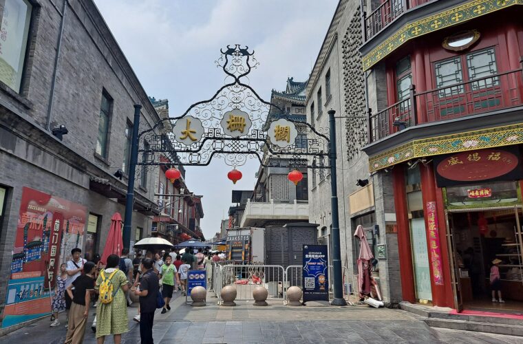 Visitors pass beneath the ornate Dashilar gateway between grey brick walls and a red traditional shopfront in Beijing.