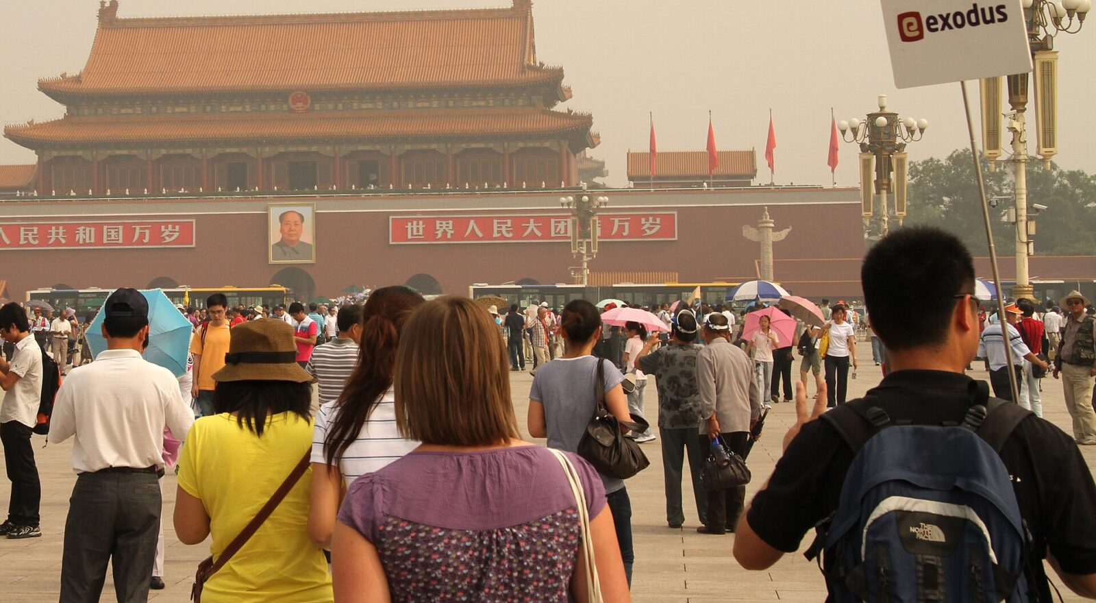 Visitors with umbrellas gather in Tiananmen Square facing the red gate, while a tour-group sign rises above the crowd.
