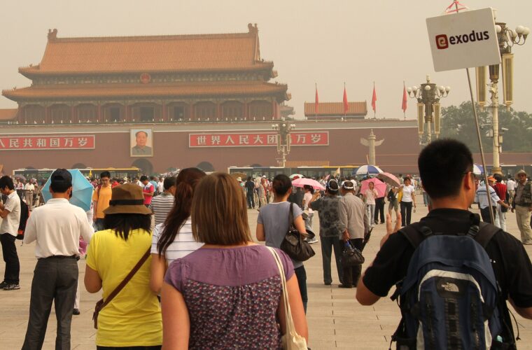 Visitors with umbrellas gather in Tiananmen Square facing the red gate, while a tour-group sign rises above the crowd.