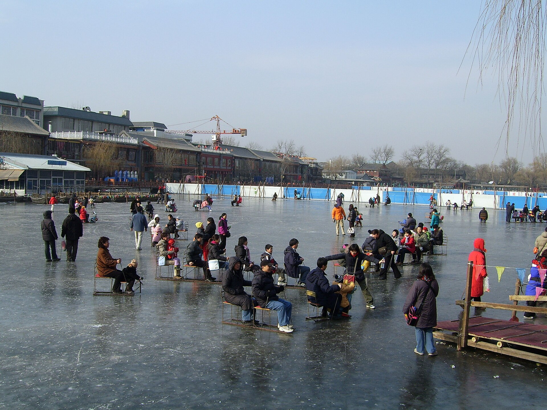 Visitors use metal ice chairs and skates on frozen Shichahai in Beijing beside low buildings and leafless willow trees.
