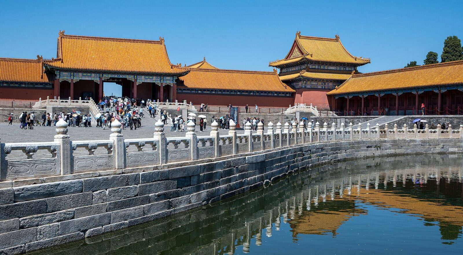 Visitors walk beside the Golden Water River in Beijing Forbidden City, where golden roofs and white balustrades reflect in the water.