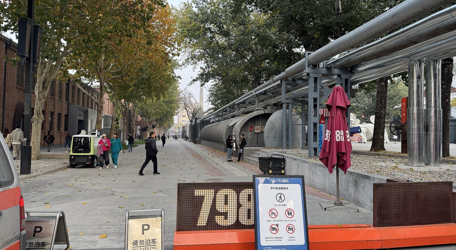 Visitors walk past brick buildings and exposed industrial pipework along a tree-lined street in Beijing's 798 Art District.