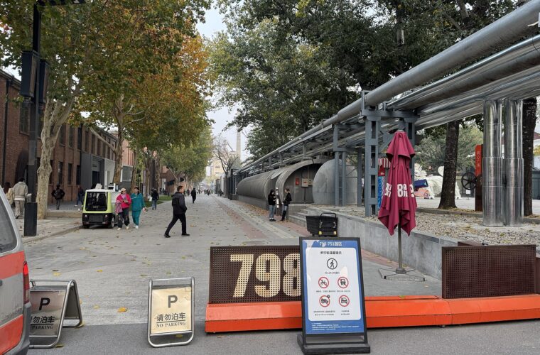 Visitors walk past brick buildings and exposed industrial pipework along a tree-lined street in Beijing's 798 Art District.