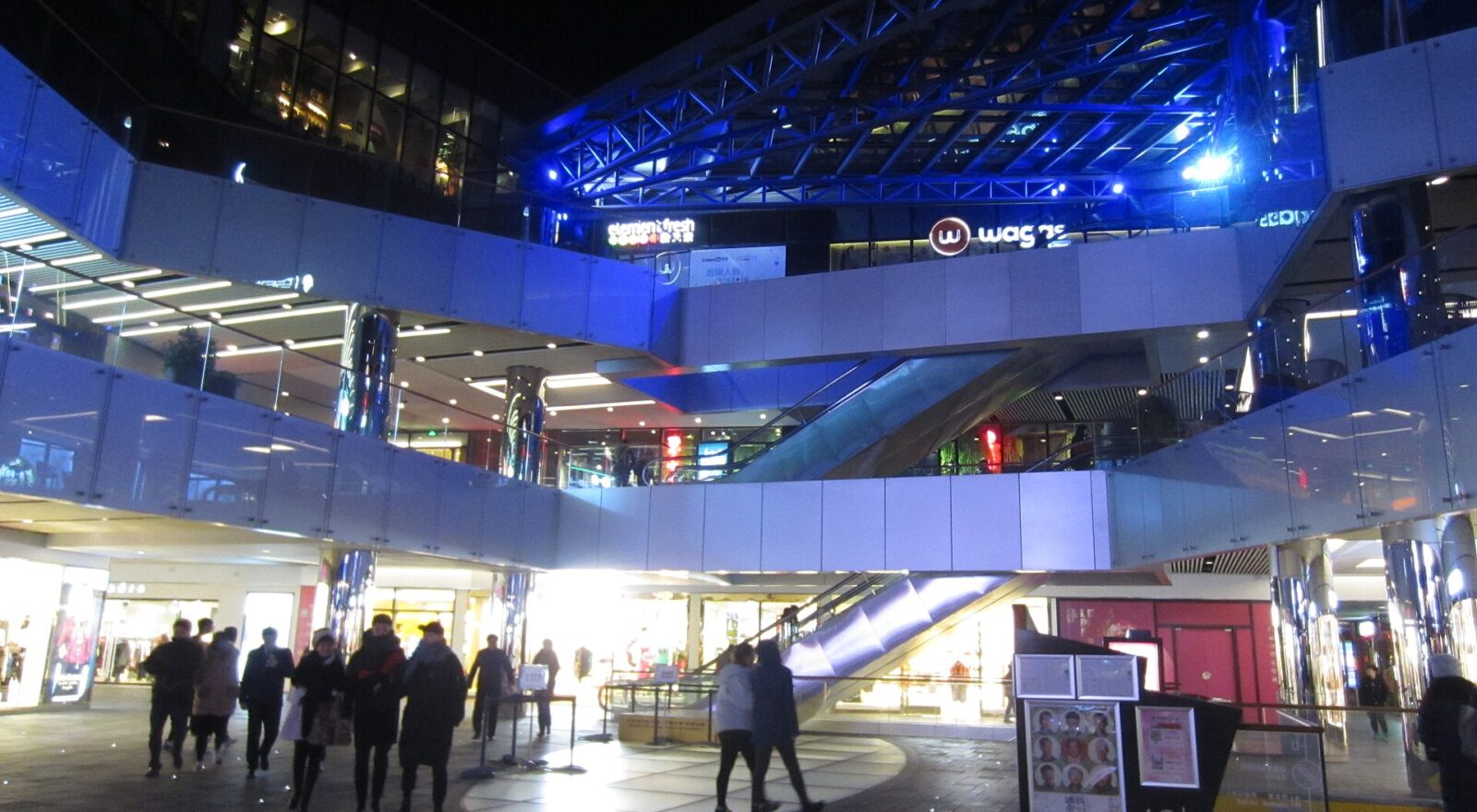 Visitors walk past illuminated shops and escalators beneath blue lighting at Taikoo Li Sanlitun in Beijing at night.