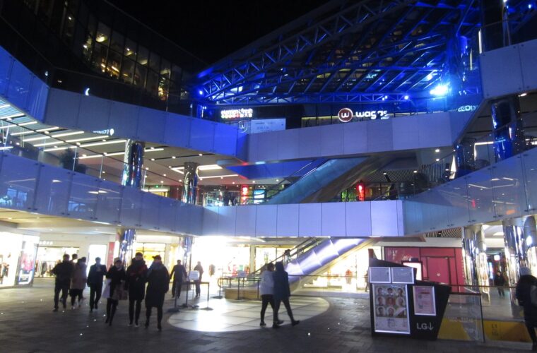 Visitors walk past illuminated shops and escalators beneath blue lighting at Taikoo Li Sanlitun in Beijing at night.