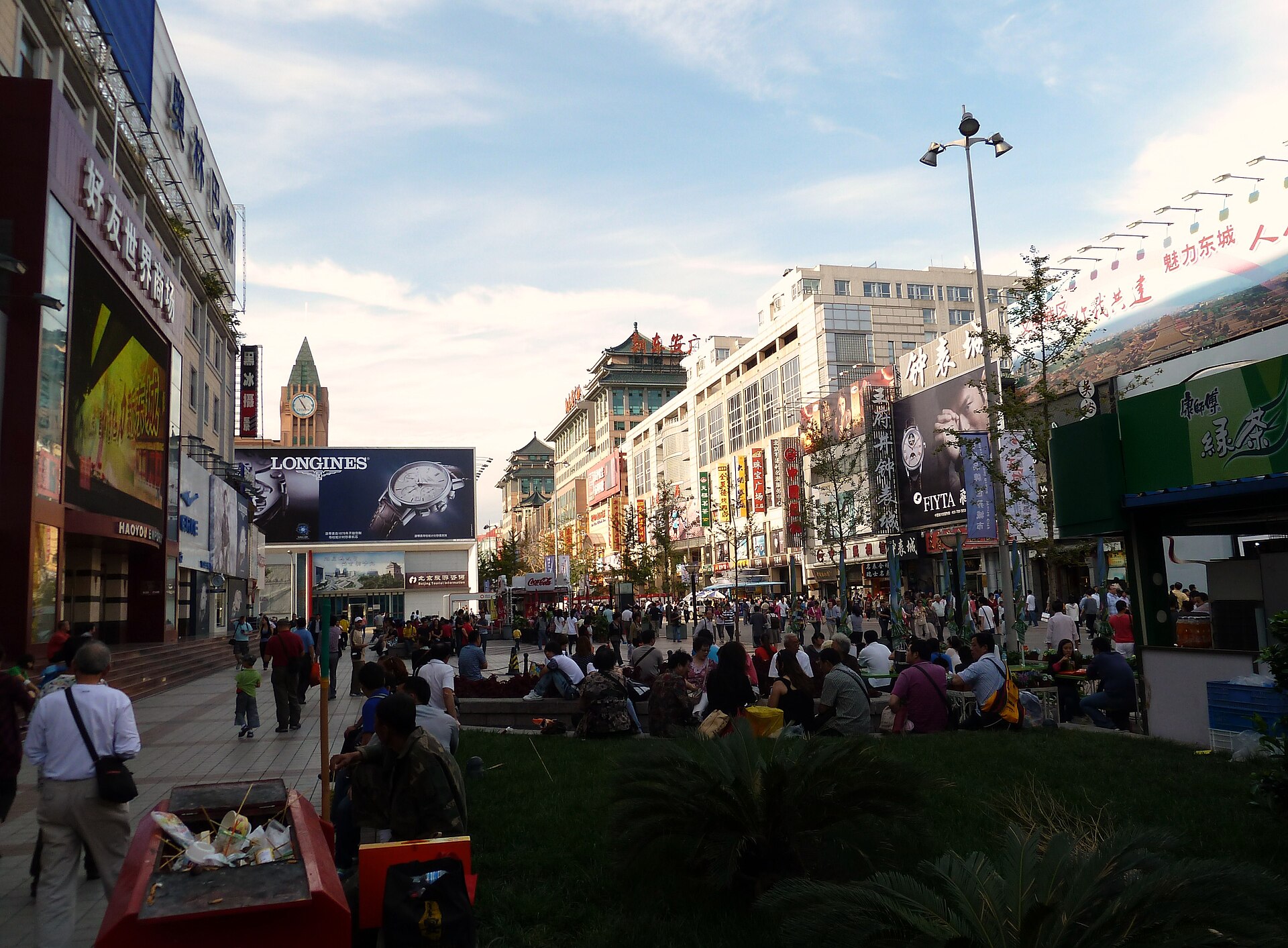 Visitors walk and sit along Beijing's busy Wangfujing pedestrian street, surrounded by department stores, advertising signs and a clock tower.