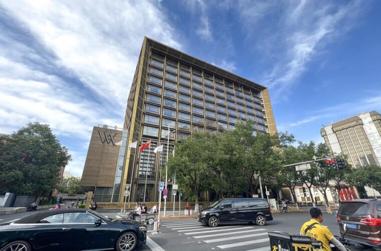 Waldorf Astoria Beijing's gold-framed facade rises above trees, flags and a busy street crossing beneath a blue sky.