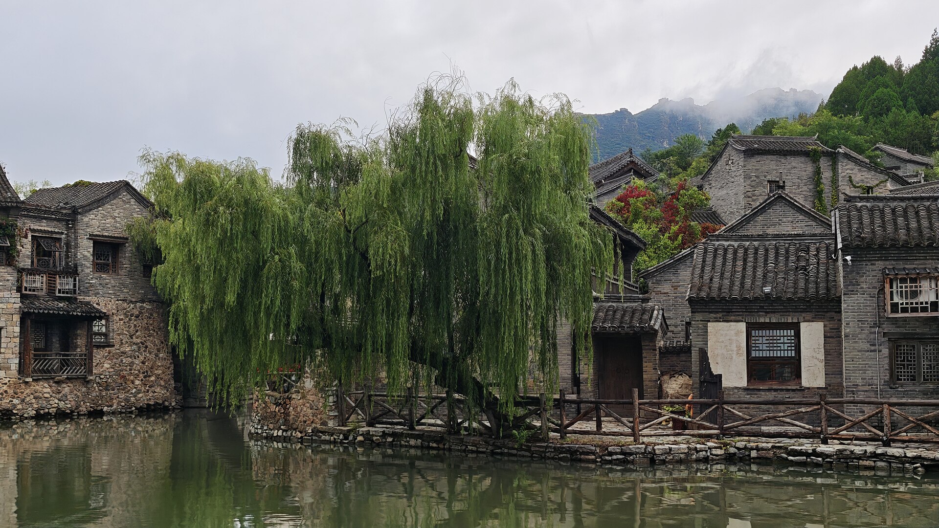 A weeping willow hangs over a canal beside grey brick houses and a wooden railing at Gubei Water Town.