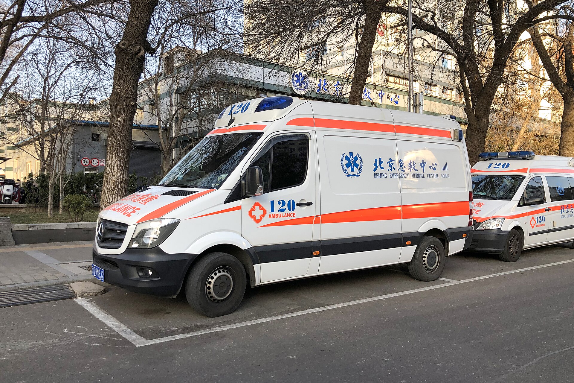 A white ambulance with orange stripes and blue 120 markings stands outside Beijing Emergency Medical Center beside another emergency vehicle.
