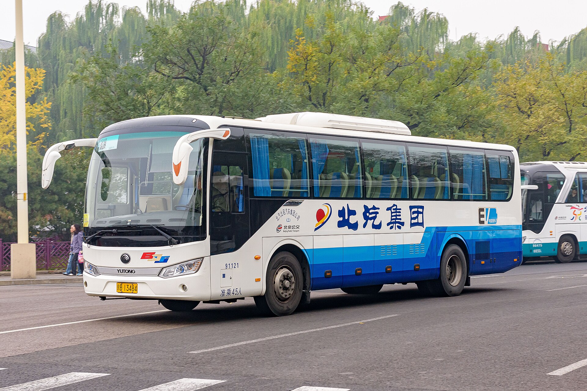 A white and blue coach bound for Mutianyu stands at Qianmen in Beijing, with willow trees behind it.