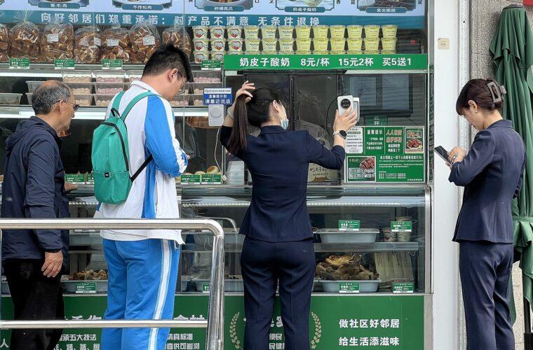 A woman seen from behind holds her phone up to a QR code reader at a Beijing food stall while other customers wait beside her