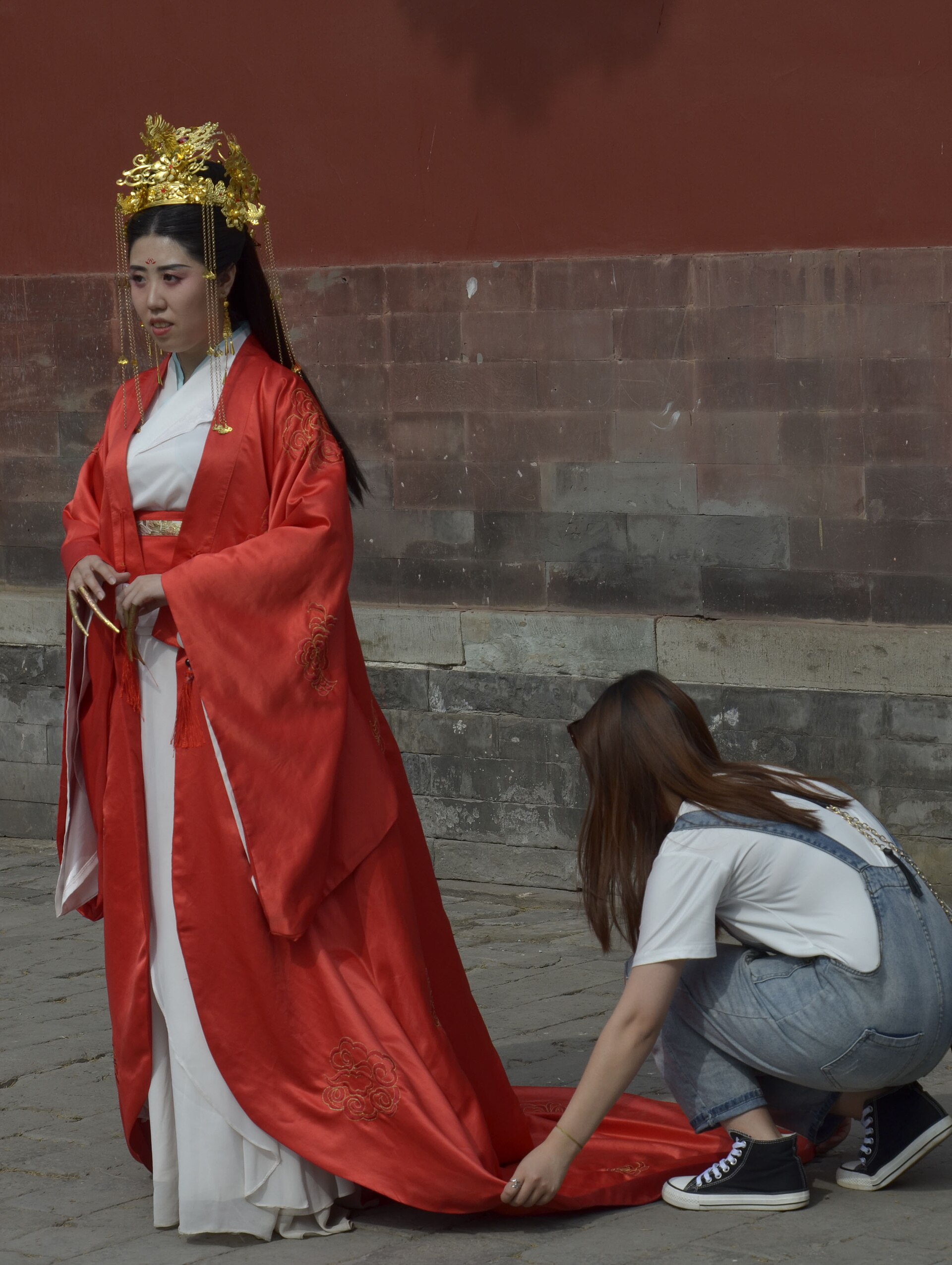A woman in a red and white hanfu with a gold headdress stands against a red palace wall in the Forbidden City while another woman arranges her train.
