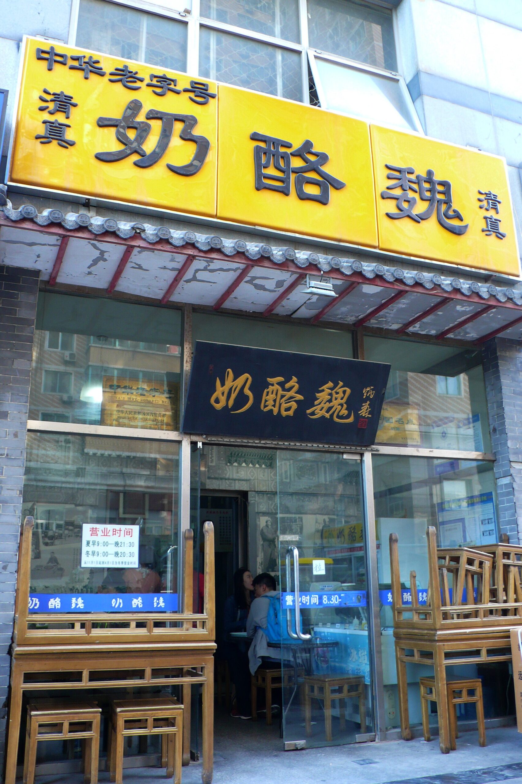 Yellow halal signage marks Wei's Yoghurt on Beijing's Niujie, above an open shop doorway with wooden stools outside.