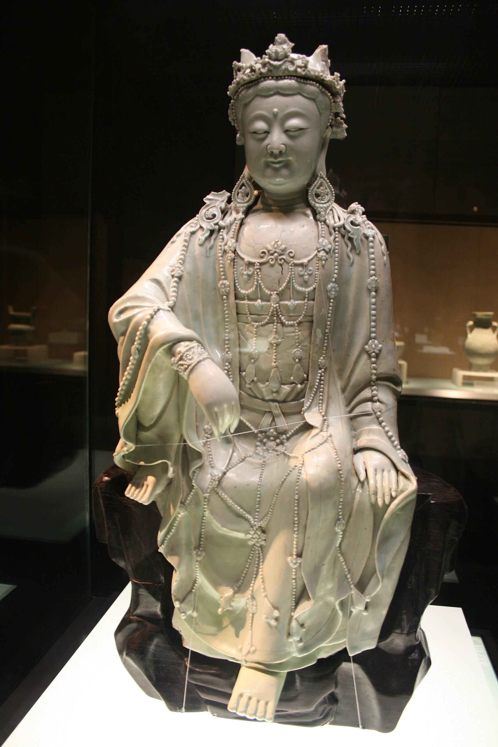A seated Yuan dynasty porcelain figure of the bodhisattva Guanyin in pale blue-white glaze, wearing a crown and strings of beads, displayed in a glass case