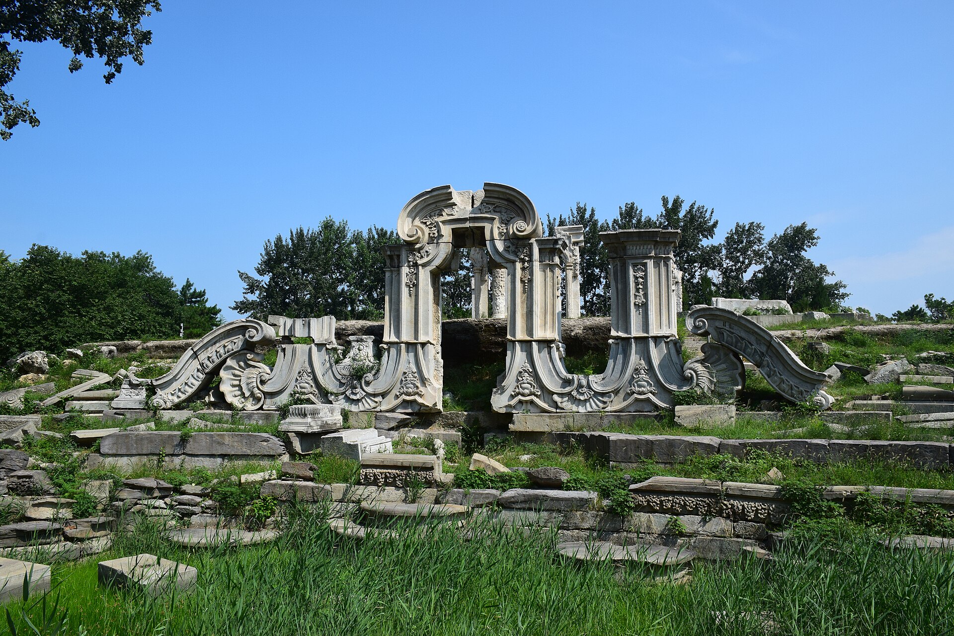 The ruined marble niche of the Great Fountain at Yuanmingyuan, carved with scrolls and shells, surrounded by fallen blocks and long grass under a blue sky